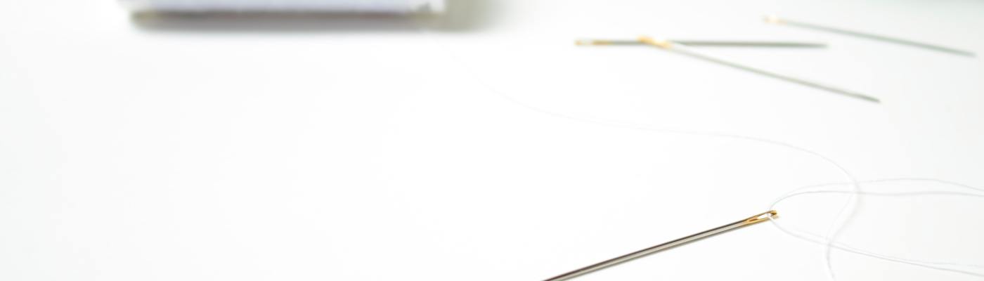 photo of a single sewing needle in foreground with spool of white thread in background